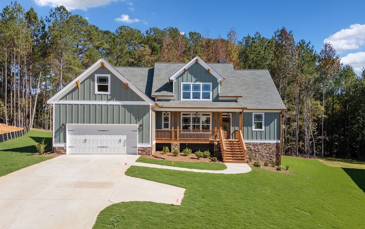 Sage green board-and-batten craftsman home with cedar front porch and stone skirt, framed by tall Georgia pines